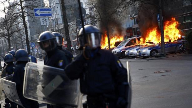 Polizisten stehen vor brennenden Polizeiautos in einer Straße.