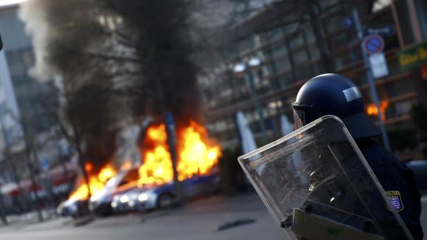 Ein Polizist mit Schutzschild steht vor brennenden Autos.