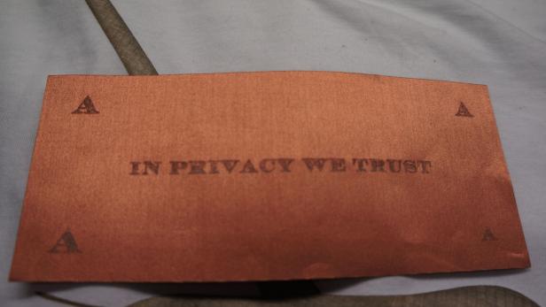 Ein braunes Stoffstück mit der Aufschrift „In Privacy We Trust“ und kleinen A-Symbolen in den Ecken.