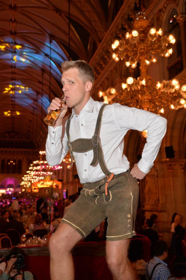 Ein Mann in Lederhosen trinkt aus einer Flasche auf einer Veranstaltung mit Kronleuchtern.