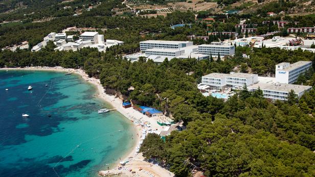 Luftaufnahme einer Küstenlinie mit türkisfarbenem Wasser, einem Strand und mehreren Hotelgebäuden.