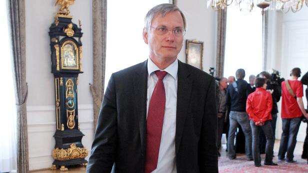 Ein Mann in Anzug und Krawatte steht in einem Raum mit einer Standuhr und anderen Personen.