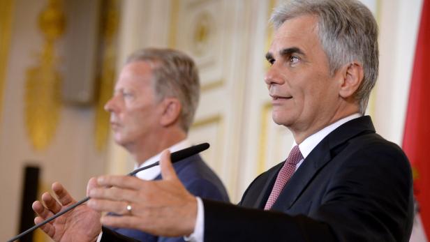 Zwei Männer in Anzügen stehen vor Mikrofonen und sprechen.