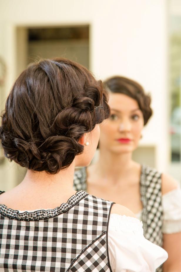 Eine Frau mit Hochsteckfrisur und Dirndl betrachtet sich im Spiegel.