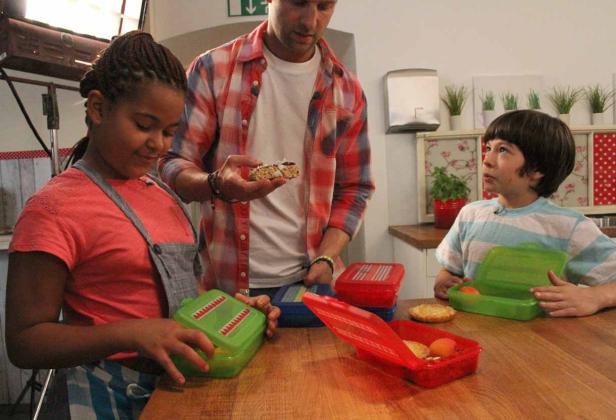 Zwei Kinder und ein Mann bereiten eine Lunchbox mit Obst und einem Müsliriegel vor.