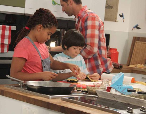 Zwei Kinder bereiten mit einem Mann in einer Küche kleine Törtchen zu.