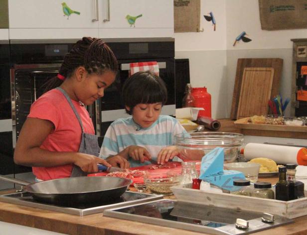 Zwei Kinder stehen in einer Küche und schneiden Zutaten für ein Gericht.