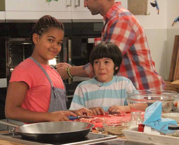 Zwei Kinder bereiten mit Hilfe eines Erwachsenen eine Mahlzeit in einer Küche zu.