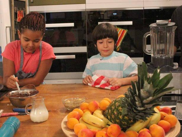 Zwei Kinder bereiten in einer Küche eine Mahlzeit mit Obst und Schokolade zu.