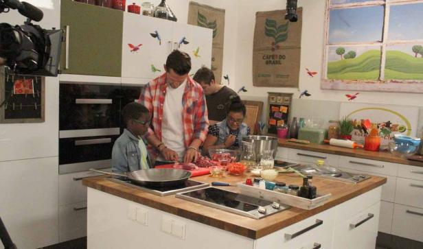 In einer Küche schneidet ein Mann Fleisch, während Kinder zusehen.