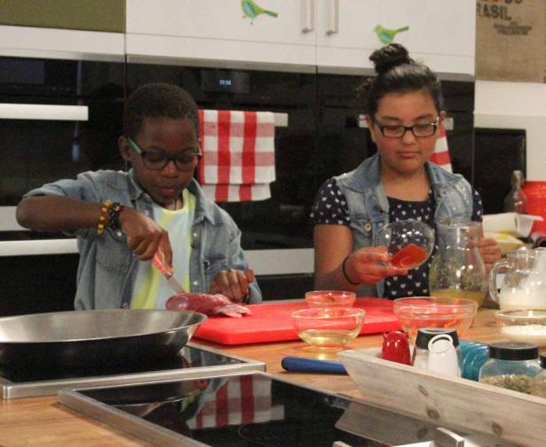 Zwei Kinder bereiten in einer Küche eine Mahlzeit zu. Ein Kind schneidet Fleisch, das andere hält eine Schüssel mit Gewürzen.