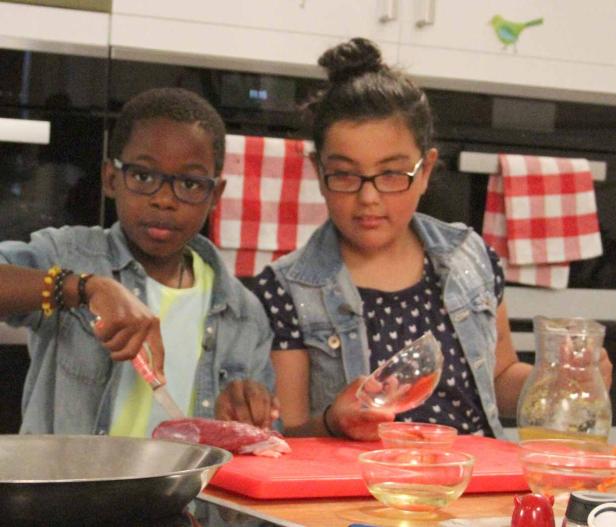 Zwei Kinder bereiten in einer Küche eine Mahlzeit zu, eines schneidet Fleisch.