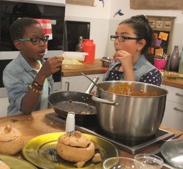 Zwei Kinder mit Brille probieren Kürbissuppe in Brottöpfen in einer Küche.