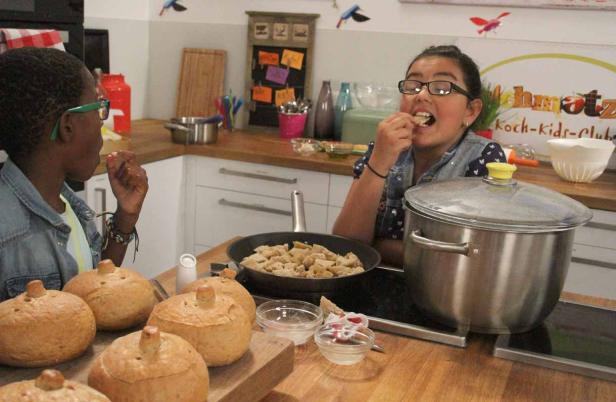 Zwei Kinder probieren in einer Küche Brot und eine Speise aus der Pfanne.