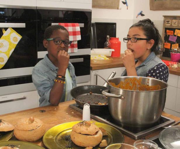 Zwei Kinder probieren Kürbissuppe in einer Küche, dekoriert für Halloween.