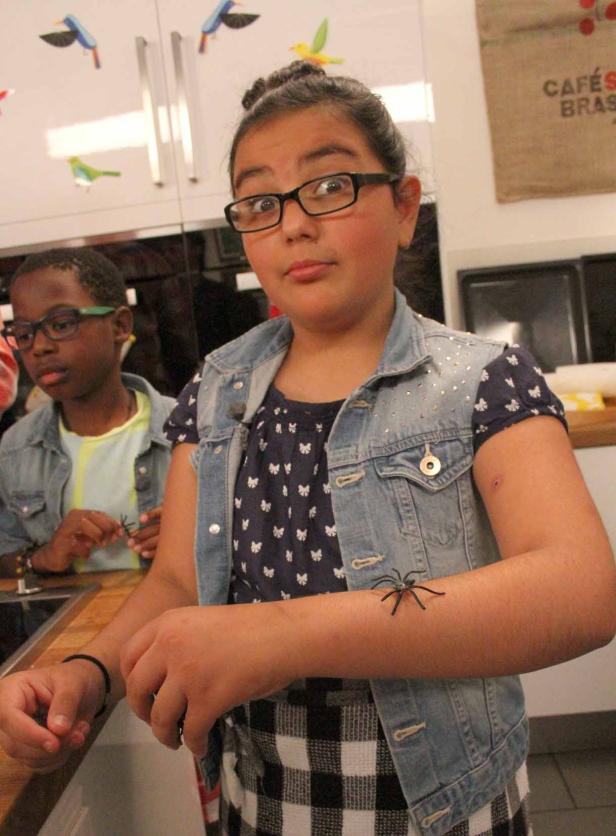 Ein Mädchen mit Brille hat eine Spielzeugspinne auf dem Arm, im Hintergrund ein Junge.