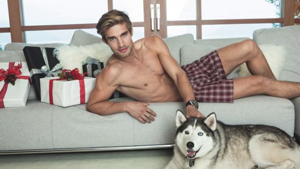 Ein Mann liegt mit einem Husky und Weihnachtsgeschenken auf einem Sofa.