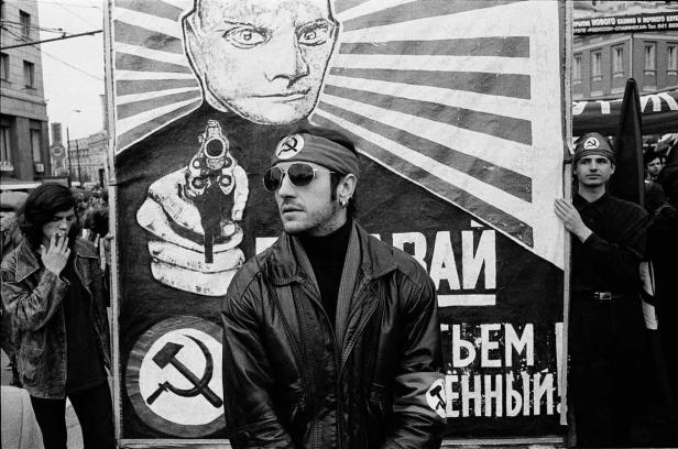 Ein Mann mit Sonnenbrille steht vor einem Plakat mit einem stilisierten Porträt und einer Pistole.