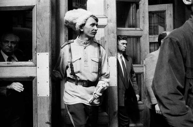 Ein Mann in traditioneller Kleidung und Pelzmütze steht vor einer Holztür.