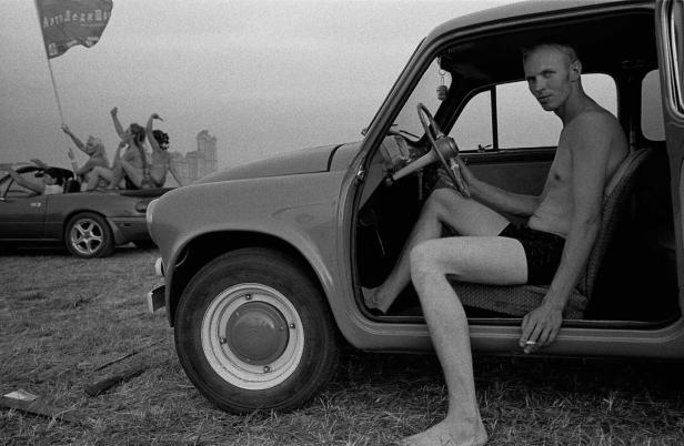 Ein barfüßiger Mann sitzt in einem alten Auto und raucht eine Zigarette. Im Hintergrund jubeln Menschen auf einem anderen Auto.