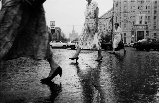 Frauen überqueren eine nasse Straße in der Stadt; im Hintergrund ist ein Wolkenkratzer zu sehen.