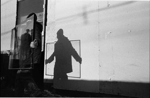 Eine Person trägt eine Glasscheibe, deren Spiegelung eine weitere Person zeigt. Der Schatten der Person ist an einer Wand zu sehen.