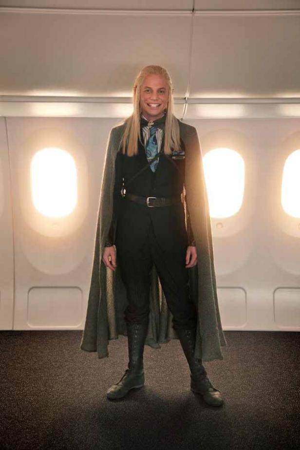 Ein Mann mit langen blonden Haaren, gekleidet wie eine Fantasy-Figur, steht in einem Flugzeug.