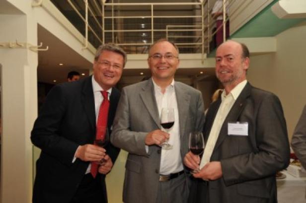 Drei Männer in Anzügen halten Weingläser in einem Innenraum.