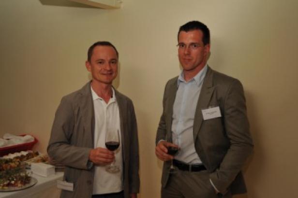 Zwei Männer in Anzügen stehen mit Weingläsern auf einer Veranstaltung.