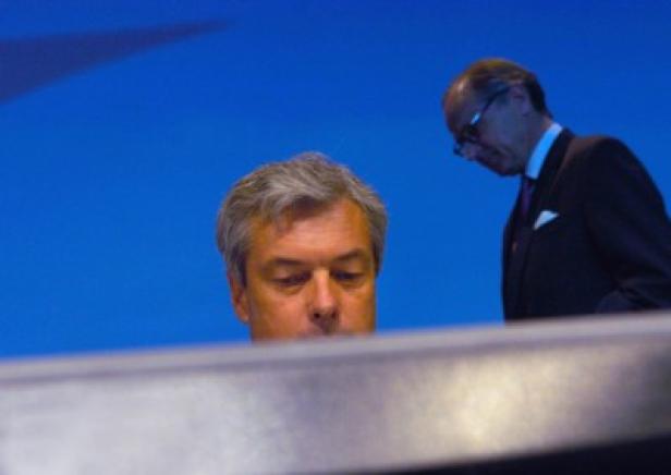 Zwei Männer in Anzügen vor einem blauen Hintergrund.