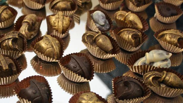 Verschiedene Pralinen in Form von Helmen und Amphoren liegen auf einer spiegelnden Oberfläche.