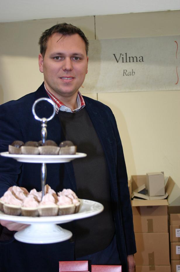Ein Mann präsentiert eine Etagere mit kleinen Kuchen vor einem Schild mit der Aufschrift „Vilma Rab“.