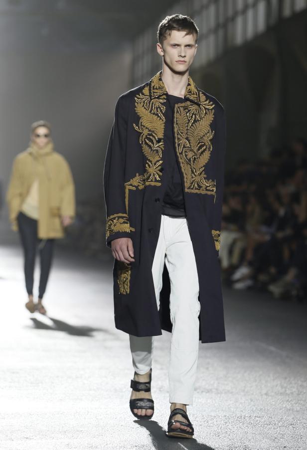 Ein Mann auf dem Laufsteg trägt einen bestickten Mantel und Sandalen.
