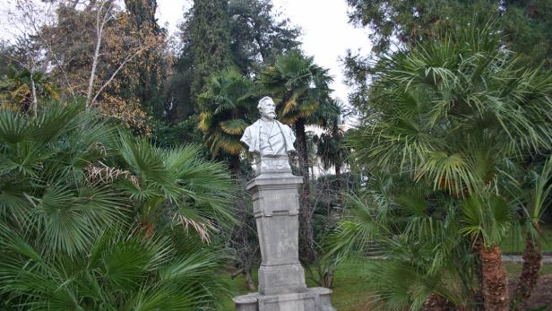 Eine Büste auf einem Sockel, umgeben von Palmen in einem Park.