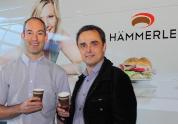 Zwei Männer halten Kaffeebecher vor einem Werbeplakat von Hämmerle.