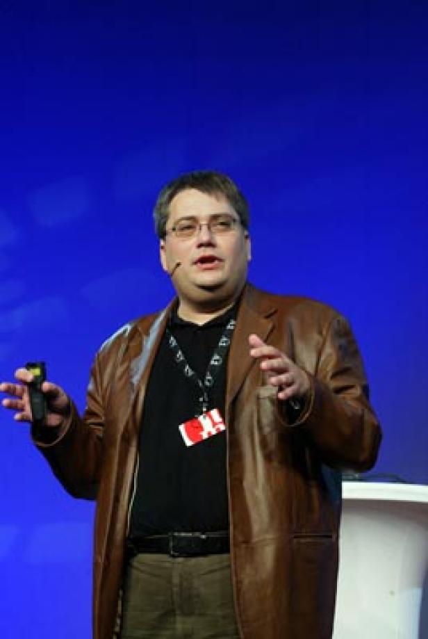 Ein Mann mit Brille und brauner Jacke hält eine Präsentation vor blauem Hintergrund.