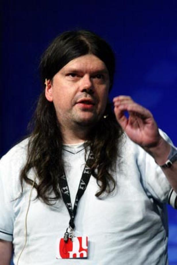 Ein Mann mit langen Haaren gestikuliert vor einem blauen Hintergrund.