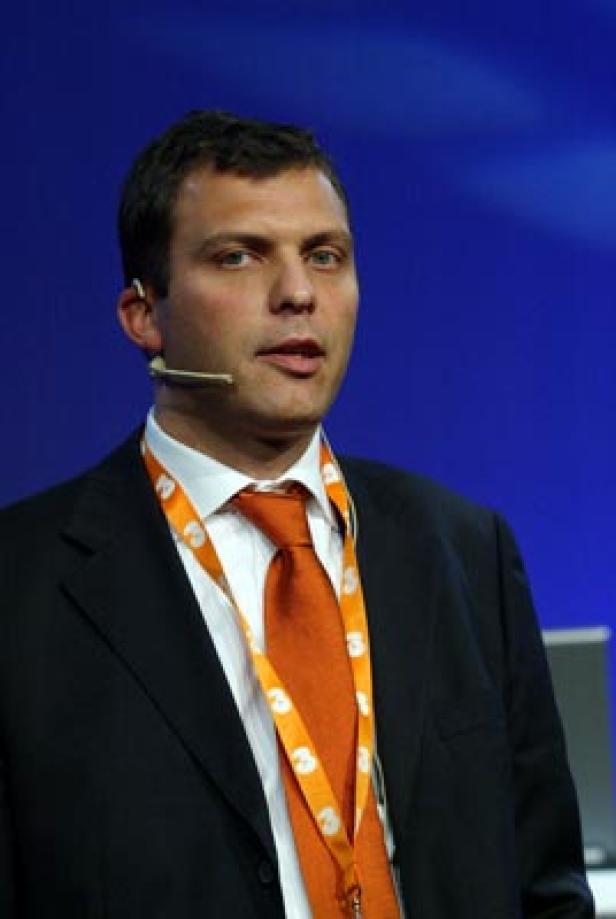 Ein Mann mit Anzug und orangefarbener Krawatte spricht in ein Mikrofon.
