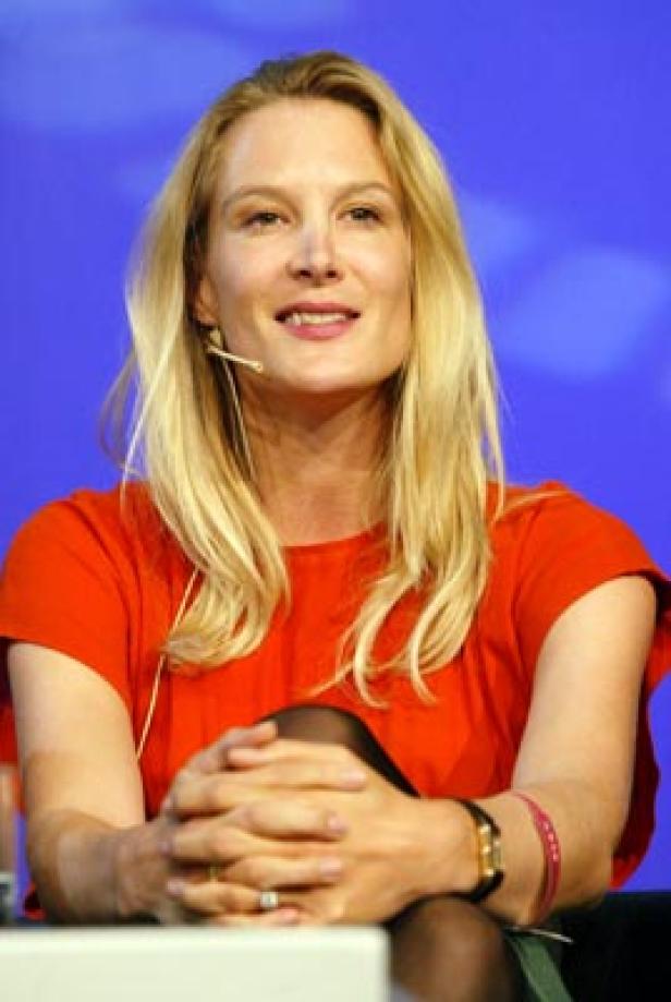 Eine blonde Frau in einem roten Kleid sitzt mit verschränkten Händen vor einem blauen Hintergrund.