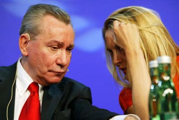 Ein Mann im Anzug und eine blonde Frau sitzen an einem Tisch vor blauem Hintergrund.