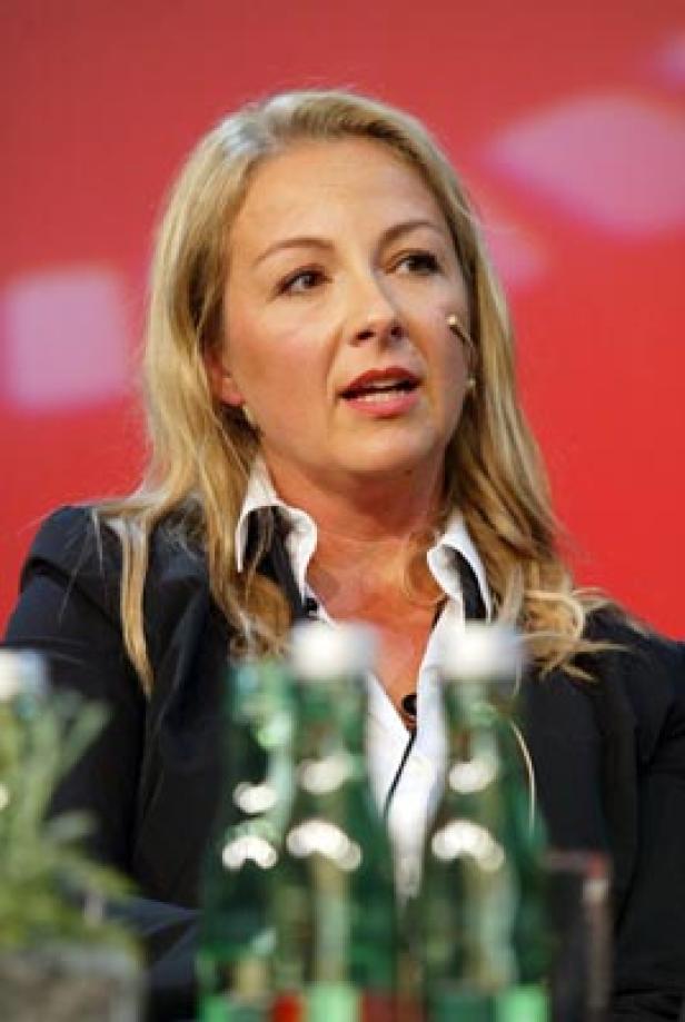 Eine blonde Frau spricht bei einer Konferenz vor einem roten Hintergrund.