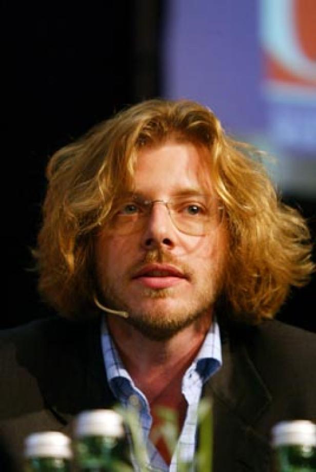 Ein Mann mit blonden, lockigen Haaren und Brille spricht in ein Mikrofon.
