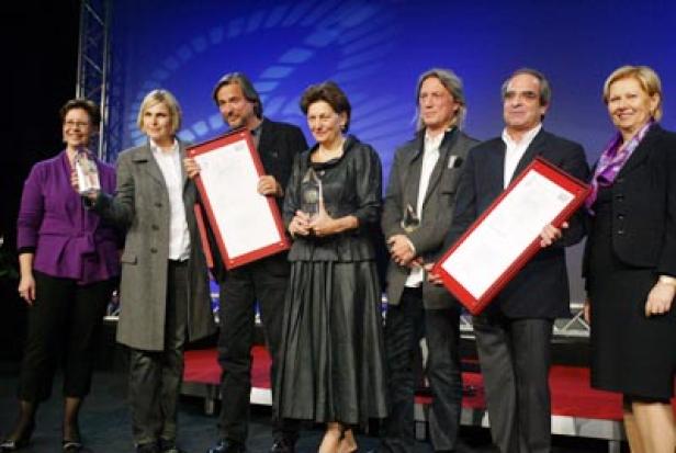 Eine Gruppe von sieben Personen steht auf einer Bühne und hält Urkunden und Trophäen.