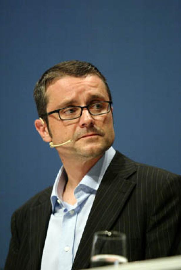 Ein Mann mit Brille und Anzug spricht vor einem blauen Hintergrund.