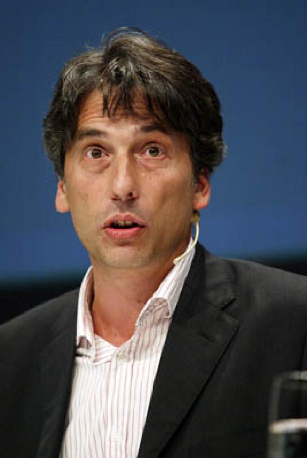 Ein Mann mit dunklen Haaren und einem Hemd mit Streifen spricht.