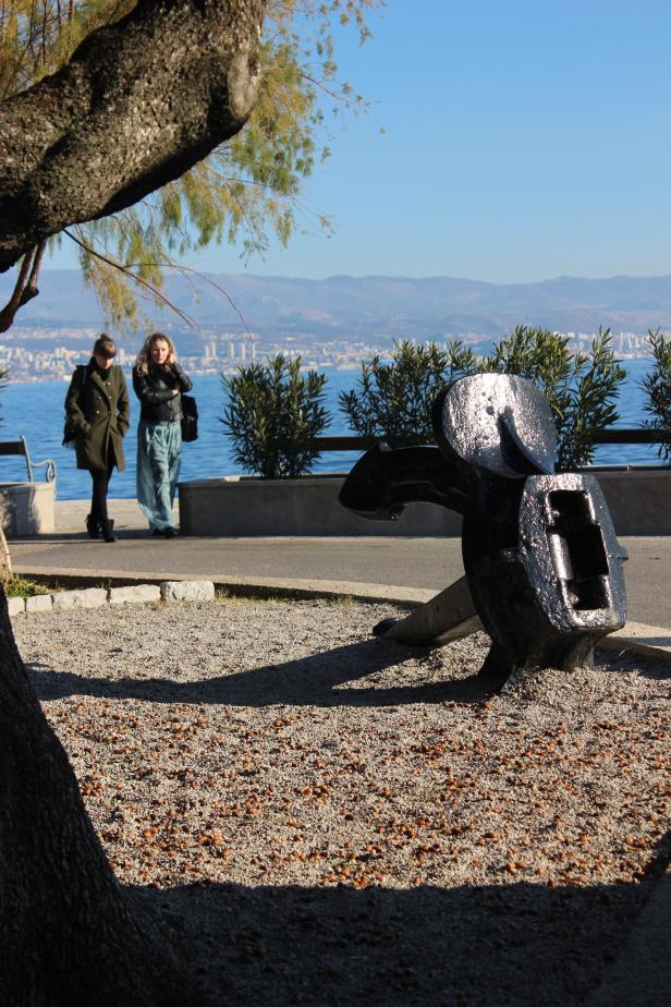 Eine alte Schiffsanker-Skulptur steht an einer Promenade mit Blick auf das Meer.