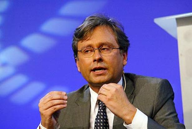 Ein Mann mit Brille und Anzug gestikuliert vor einem blauen Hintergrund.