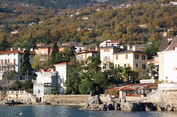 Häuser mit roten Dächern an der Küste mit einem bewaldeten Hügel im Hintergrund.