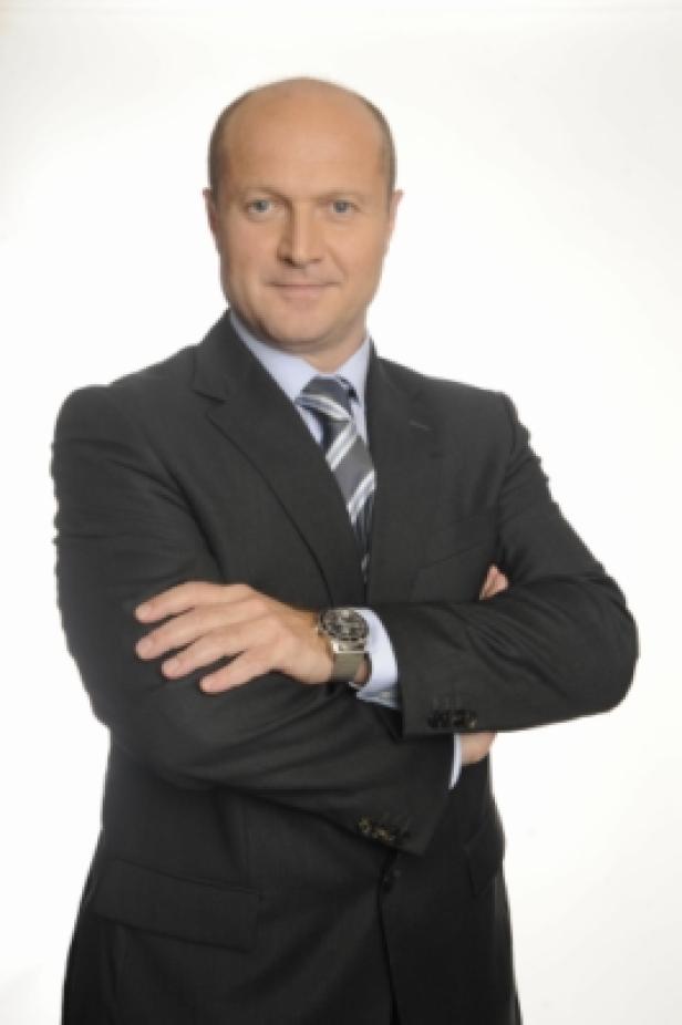 Ein Mann im Anzug mit verschränkten Armen vor einem weißen Hintergrund.