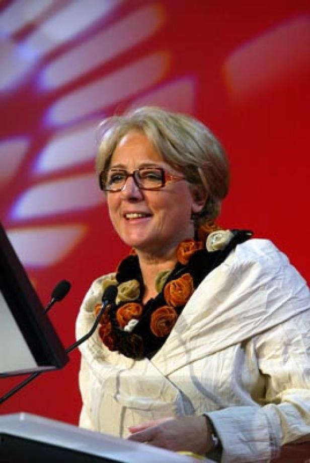 Eine Frau mit Brille hält eine Rede vor einem roten Hintergrund.
